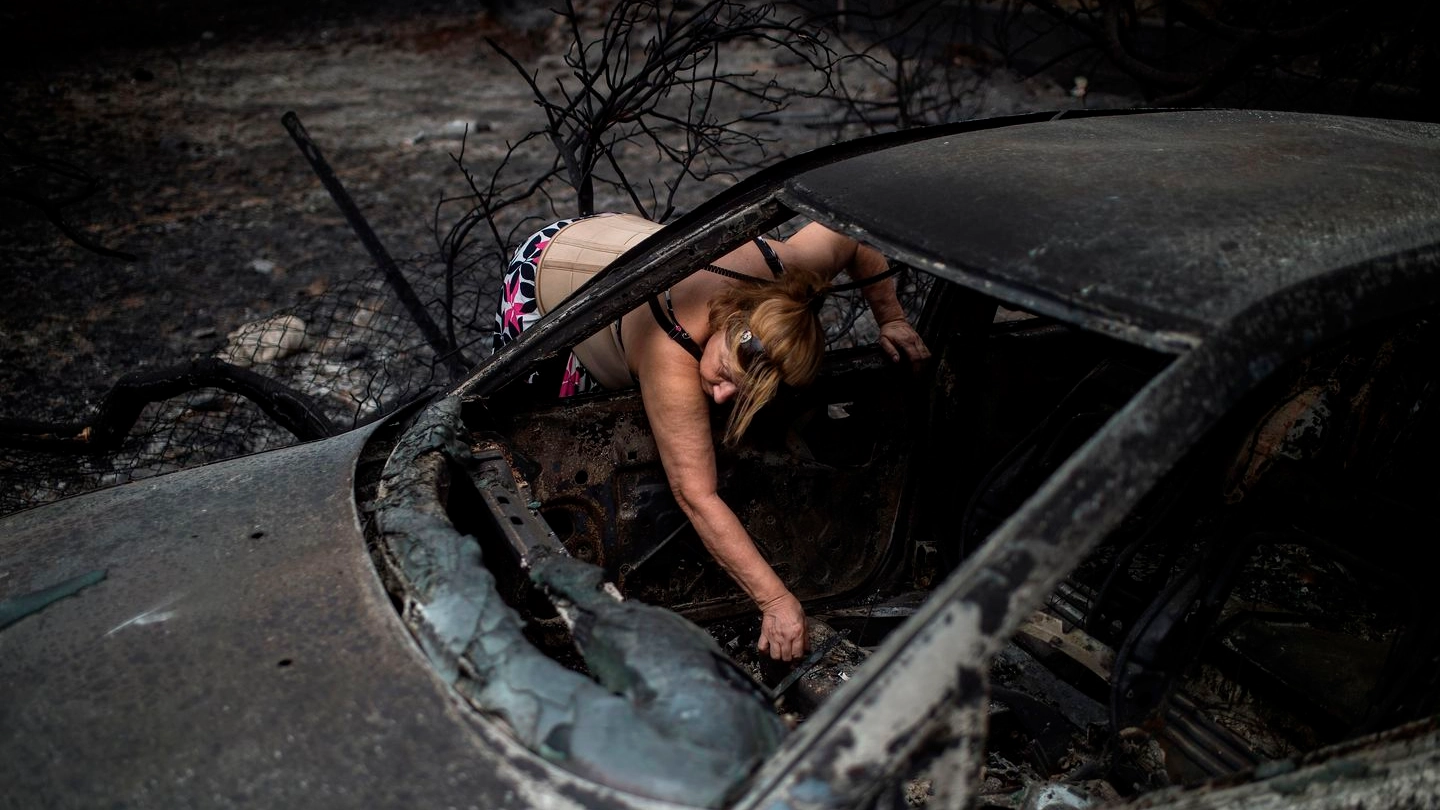 Grecia, una donna cerca di trovare dei fogli nell'auto carbonizzata a Mati (LaPresse)