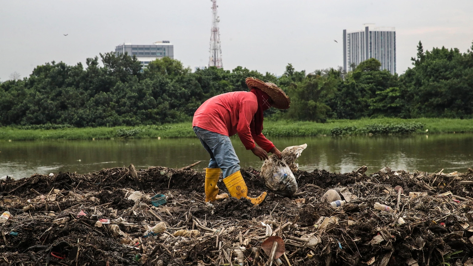 Un uomo raccoglie rifiuti lungo la riva di un fiume (Ansa)