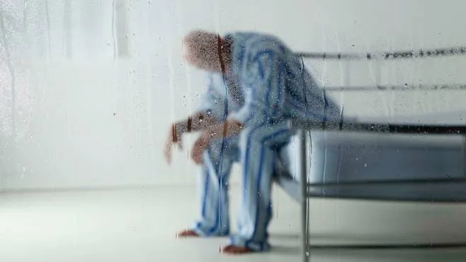 Shot of a man - possibly a hospital patient or inmate - sitting on his bed in his striped pyjamas. Seen through a rain-spattered window - focus on window.