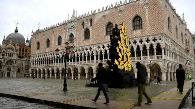People walk on elevated planks across a flooded St. Mark's Square past the Doge's Palace (Rear) and a luminous Christmas Tree installation by Italian artist Fabrizio Plessi (Front C) on December 8, 2020 following a high tide "Alta Acqua" event following heavy rains and strong winds, and the mobile gates of the MOSE Experimental Electromechanical Module that protects the city of Venice from floods, were not lifted (Photo by ANDREA PATTARO / AFP) / RESTRICTED TO EDITORIAL USE - MANDATORY MENTION OF THE ARTIST UPON PUBLICATION - TO ILLUSTRATE THE EVENT AS SPECIFIED IN THE CAPTION