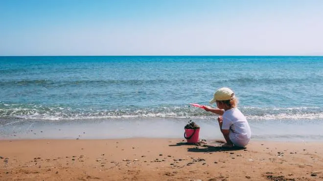 A child plays with a pink plastic bucket and shovel in the sand on a beach, close to the sea. Crete, Greece.