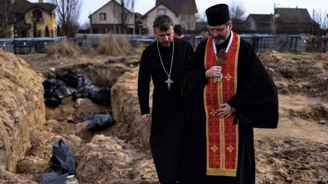 Priests pray at body bags in a mass grave in the garden surrounding the St Andrew church in Bucha, on April 7, 2022, amid Russia's military invasion launched on Ukraine. - The UN humanitarian chief said on April 7 during a visit to the town of Bucha outside the Ukrainian capital Kyiv, which included a stop at the site of a mass grave that Ukrainians had dug near a church, that investigators would probe civilian deaths uncovered after Russian troops withdrew. (Photo by RONALDO SCHEMIDT / AFP)
