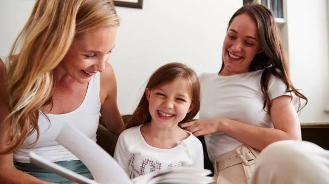 Same Sex Female Couple Reading Book With Daughter At Home Together Whilst Tickling Her