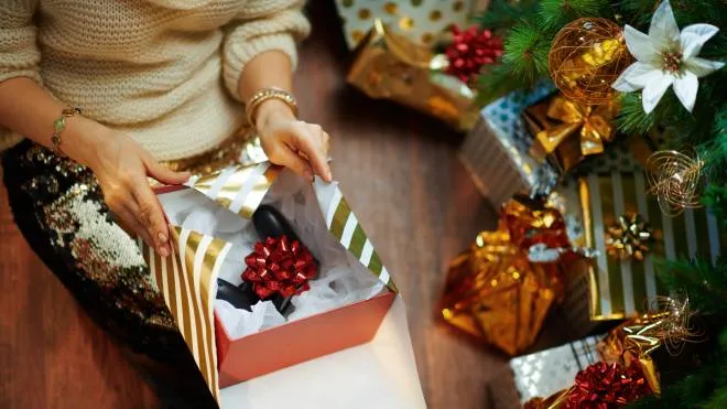 Closeup on trendy woman in gold sequin skirt and white sweater under decorated Christmas tree near present boxes wrapping Christmas gift joystick for him.
