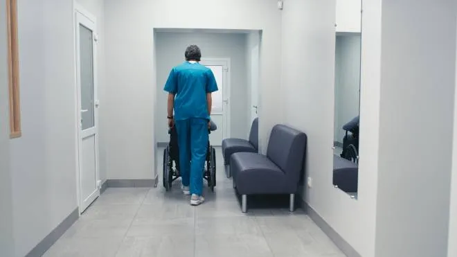 Back view of man in scrubs pushing wheelchair with handicapped male patient while working in corridor of modern clinic