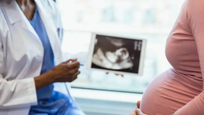 A photo with the focus on the unrecognizable pregnant woman in the foreground as the unrecognizable doctor shows her an ultrasound on a digital tablet in the background.