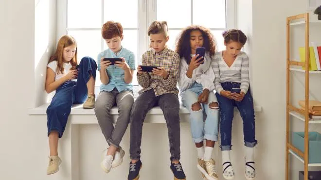 Group of children of different nationalities sit in a row on the windowsill and with lack of emotion play online games or read social networks on mobile phones. Technologies that spoil childhood.