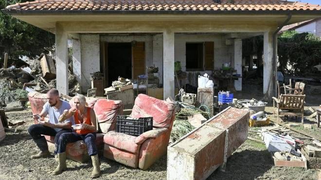 Una coppia mangia sul divano distrutto posizionato in giardino dopo l'alluvione (Ansa)